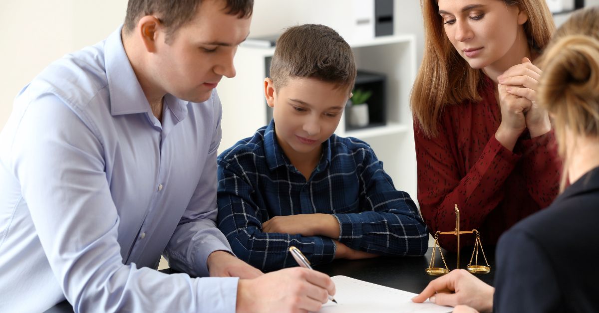 A family, including a child, sits at a table with a professional. The man signs a document as others watch, with a scale of justice visible on the table.