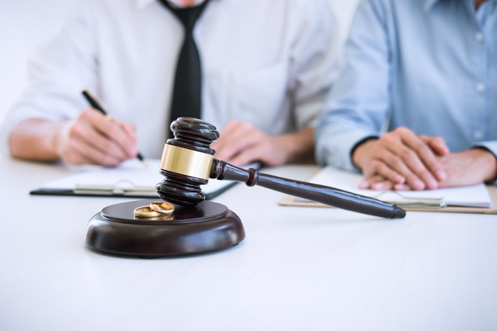 A judge's gavel rests on a sound block with two wedding rings. Two blurred individuals review documents in the background, suggesting a legal or divorce proceeding.