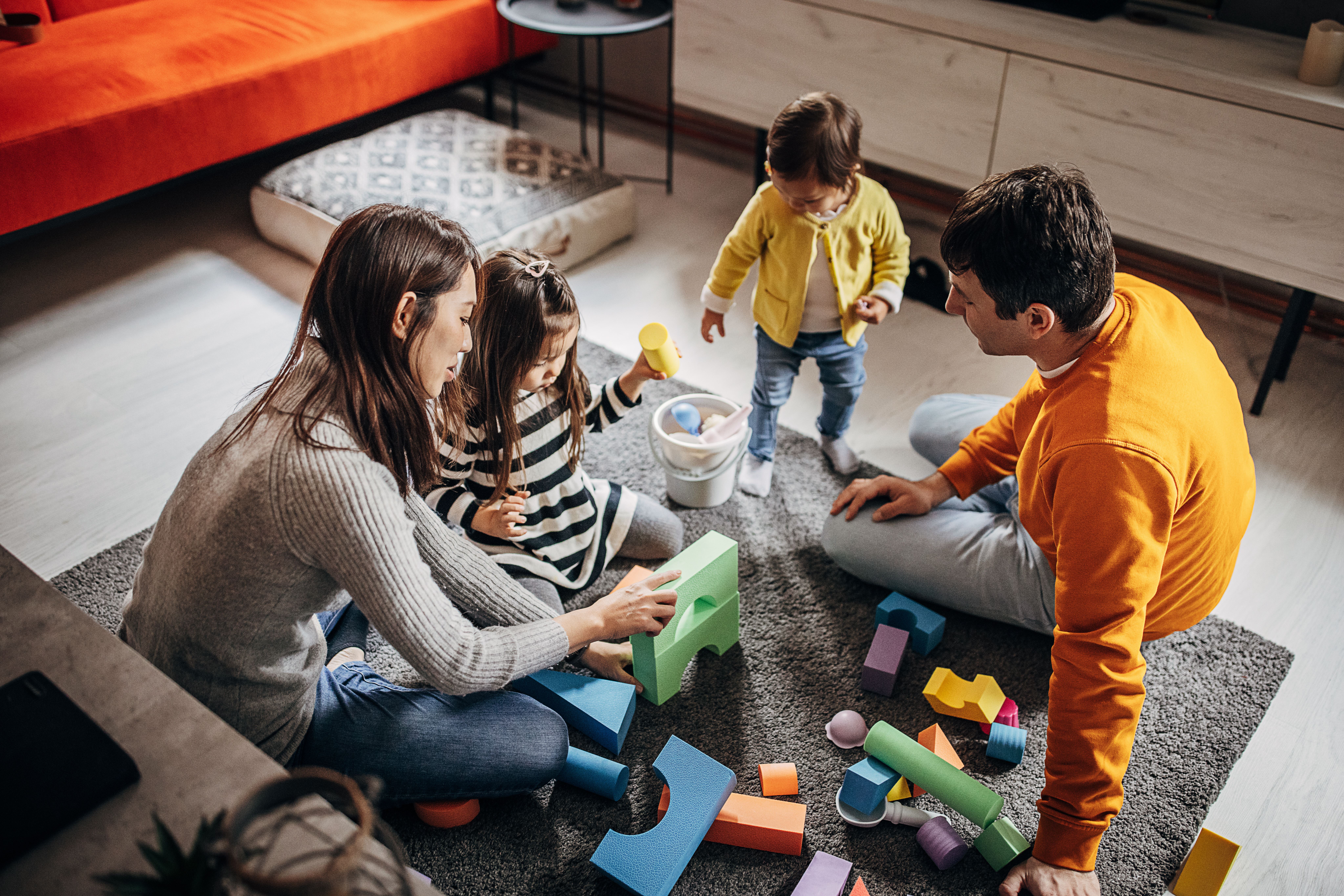 A happy family plays with colorful building blocks on a rug. Two parents interact with their two young children in a cozy living room.