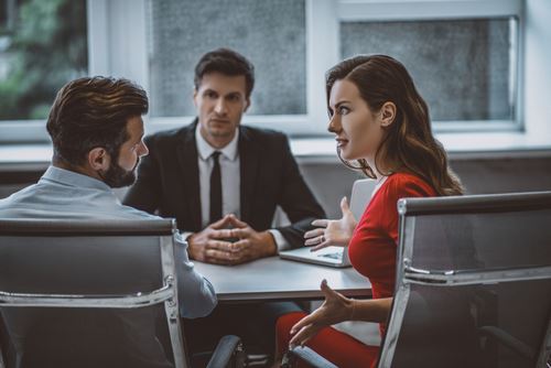 A woman in a red dress speaks animatedly to a man across a table, while a person in a suit sits between them, observing. This image depicts a meeting or mediation setting.